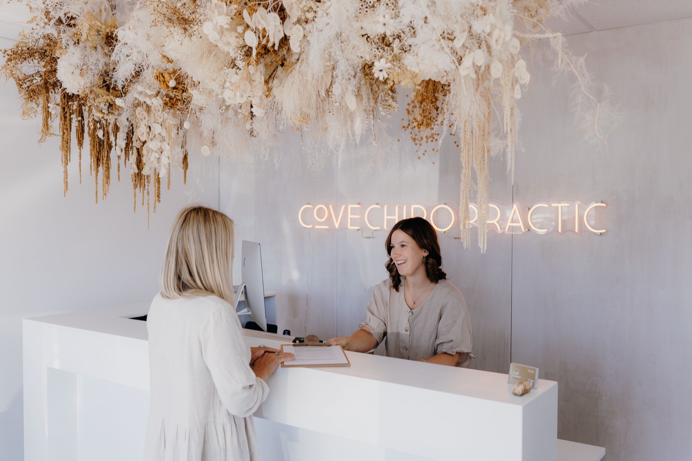 The welcoming reception at Cove Chiropractic, One Tree Point — bright, warm clinic with dried floral arrangement and neon sign